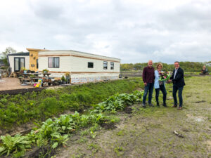 Anja Mellema en Johannes Scheerhoorn ontvangen een bloemetje uit handen van wethouder Hans Haze. Op de achtergrond de caravan waar ze tijdelijk in wonen.